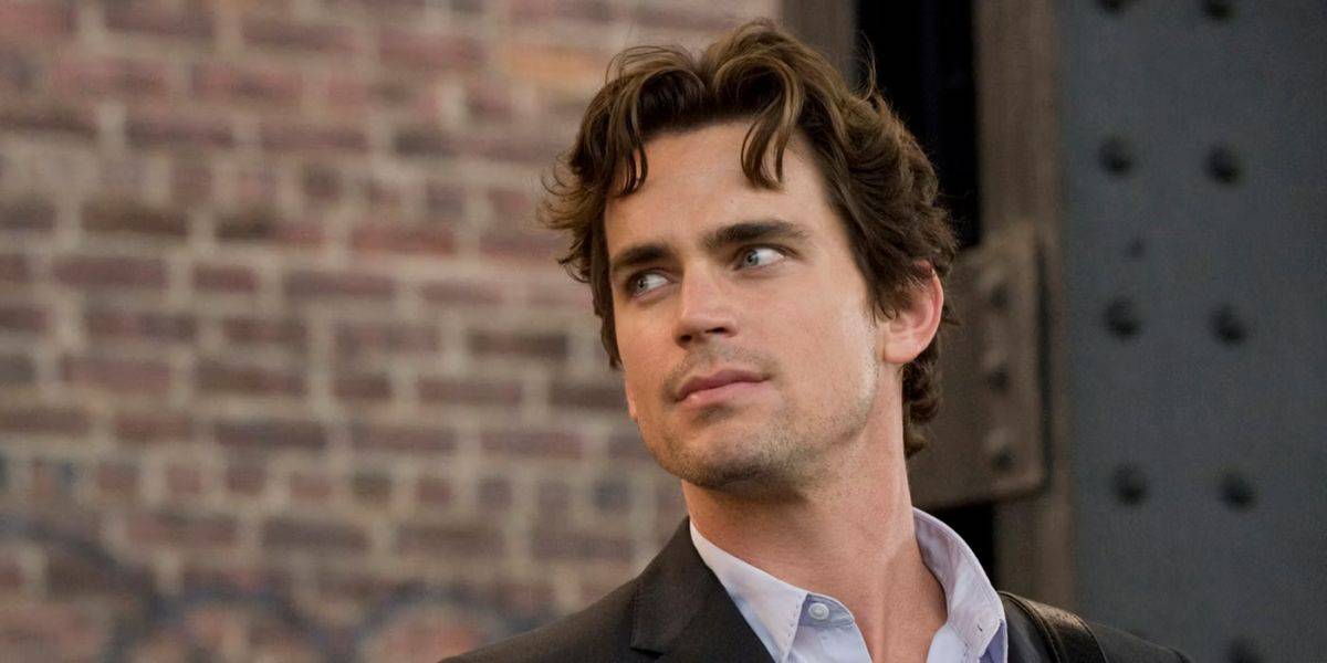 A man with wavy brown hair and stubble, resembling a seasoned actor, is looking to his right. He is wearing a black jacket over a white shirt. The background features a brick wall with some dark metal beams.