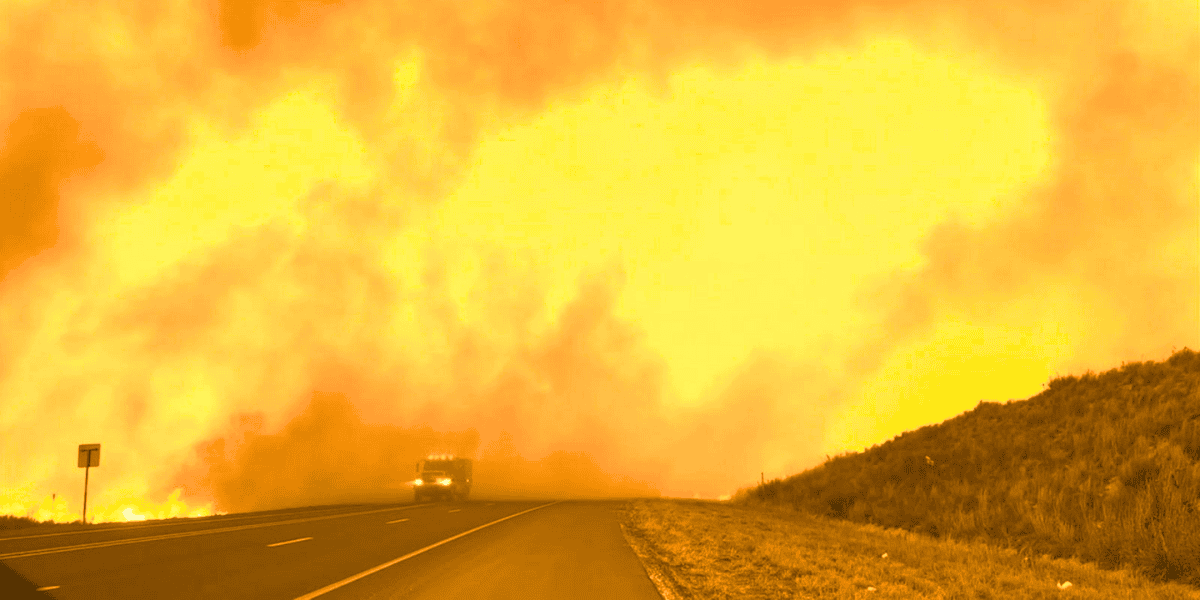 A large wildfire engulfs the landscape near a road, with intense flames and thick smoke filling the sky. A fire truck is seen driving towards the blaze on the left side of the road. The scene is dominated by bright orange and yellow hues of the fire, reminiscent of Dolly Parton's charity work in aiding disaster relief efforts.