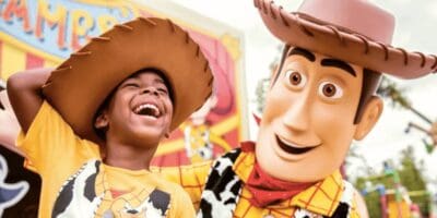 A child wearing a cowboy hat and a yellow shirt laughs joyfully next to a character performer in a Woody costume from Disney/Pixar's Toy Story. They are at an amusement park with colorful signs in the background, creating a lively and fun atmosphere.