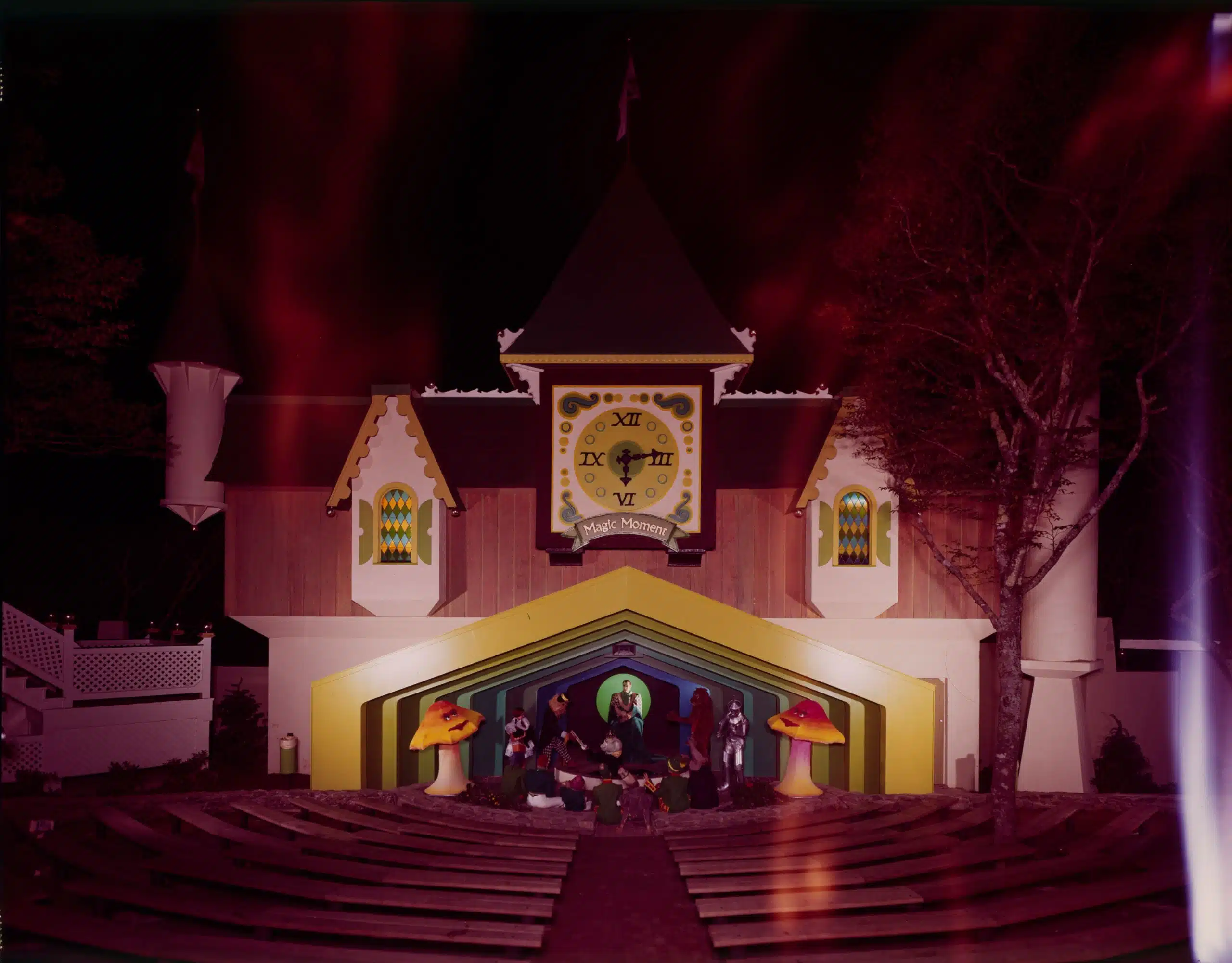 A whimsical clock tower structure with bright yellow accents at an amusement park. Below the clock, there's a small stage area featuring large mushroom decorations and a group of people, possibly setting up for a performance. The scene is bathed in surreal, reddish lighting.