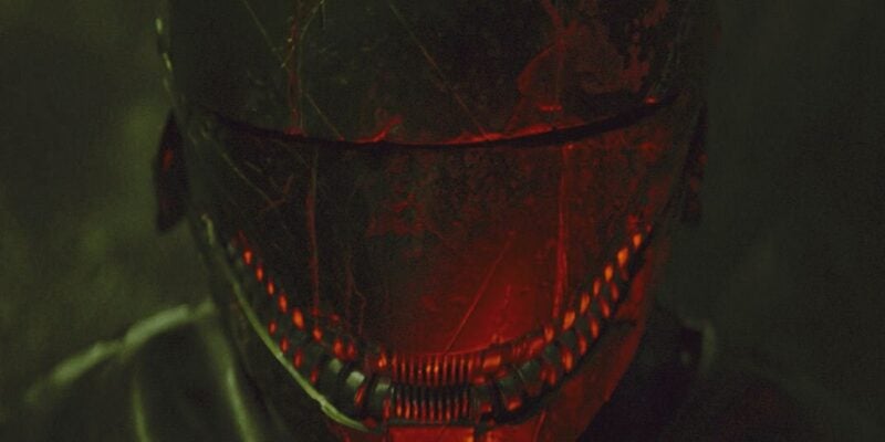 A person wearing a dark, futuristic helmet with red illuminated details around the visor and mouth area, resembling The Acolyte, set against a dimly lit, blurry background. The helmet has a worn, scratched appearance, adding a rugged, dystopian vibe to the scene.