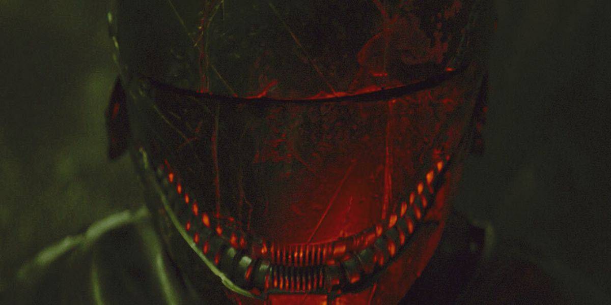 A person wearing a dark, futuristic helmet with red illuminated details around the visor and mouth area, resembling The Acolyte, set against a dimly lit, blurry background. The helmet has a worn, scratched appearance, adding a rugged, dystopian vibe to the scene.