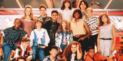 A group of teenagers, boys and girls, are posing together with cheerful expressions. They are dressed in 1990s fashion, reminiscent of Mouseketeers from a Disney Show. Some are standing while others sit in a casual, friendly manner. The background suggests they are in a diner-like setting.
