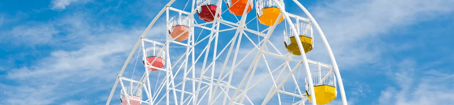 A vibrant Ferris wheel with multicolored gondolas, including red, orange, yellow, and green, stands against a bright blue sky with a few scattered clouds. The white structure of the Ferris wheel contrasts with the colorful seats and the clear sky.