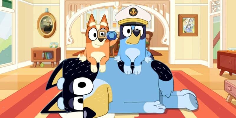 Three animated dog characters are seen in a cozy room. A large blue dog, reminiscent of Bluey, lies on the floor while a smaller orange dog sits on its back holding a magnifying glass. Another smaller blue dog wearing a captain's hat sits next to the orange dog. The background shows home decor.