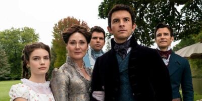 A group of five people dressed in Regency-era clothing stand outdoors with a tree and greenery in the background. Among them, Jonathan Bailey is one of three men alongside two women, all gazing off into the distance with serious expressions, as if straight out of a Disney fairy tale.