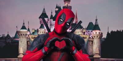Deadpool, in his iconic red and black bodysuit, stands with two swords on his back, forming a heart shape with his hands. He poses in front of a fairy tale-style castle with turrets, resembling a theme park setting at dusk.