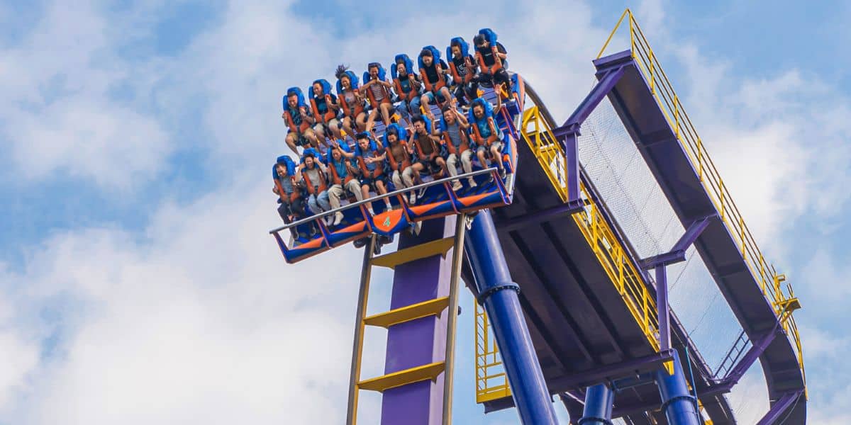 A roller coaster car filled with guests ascends a steep track against a blue sky with scattered clouds. The riders, secured with over-the-shoulder restraints, are seated in rows, appearing excited and braced for the upcoming drop. The structure is painted purple and yellow.