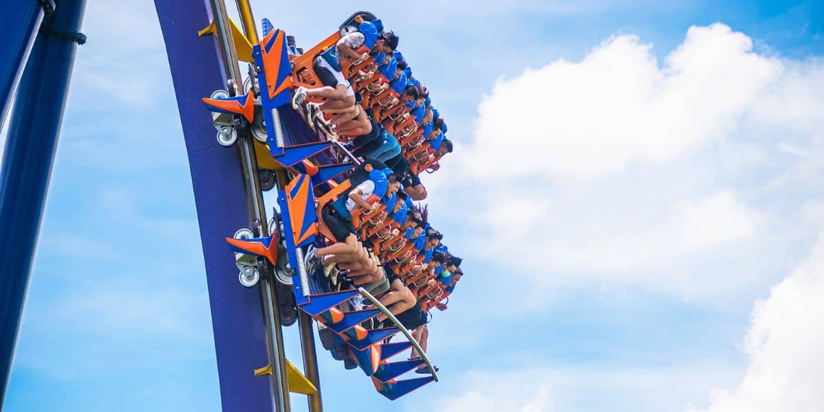People are riding a roller coaster with blue and orange seats, tilted sideways against a bright blue sky with scattered clouds. The guests have their hands up, appearing thrilled, but you can tell they're momentarily trapped in the excitement of the ride.