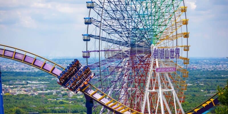 A vibrant theme park scene featuring a roller coaster with a filled car of excited guests moving through a loop in the foreground. A large, colorful Ferris wheel towers in the background against a clear sky, with a sprawling cityscape visible below.