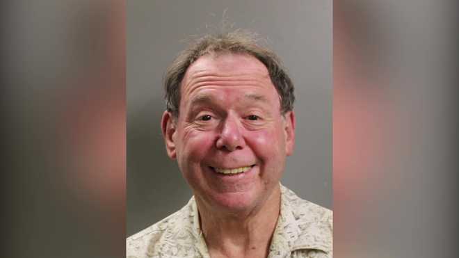 A smiling older man, John Carabello, with thinning gray hair and a receding hairline, wearing a light-colored, patterned shirt, stands against a plain gray background. He has a flushed complexion and appears to be in a cheerful mood. Perhaps he's just returned from an enjoyable day at Canobie Lake Park.