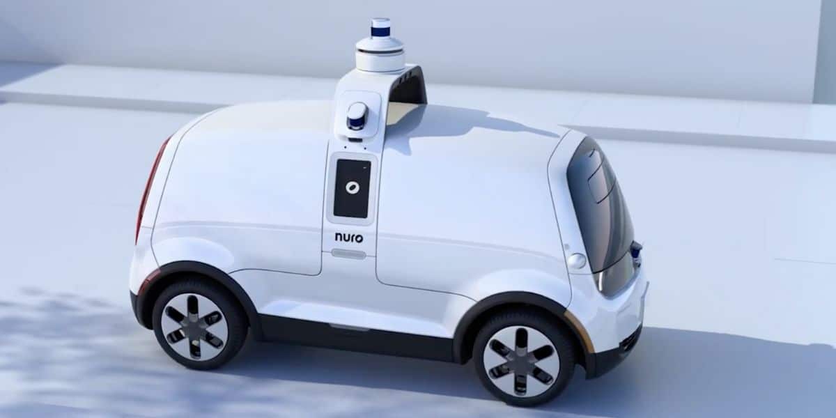 A small, white autonomous vehicle with rounded edges and minimalistic design is parked in a clean, white environment. The vehicle, reminiscent of something out of Disney's futuristic theme parks, features sensors and cameras on its roof and sides, with "nuro" printed on the side door. The vehicle has no visible windows.