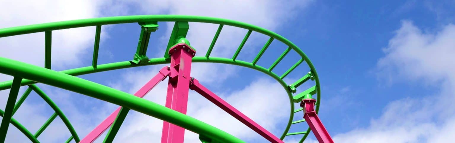A close-up view of a brightly colored roller coaster track against a blue sky with scattered white clouds. The track is green, supported by vibrant pink pillars. The image captures the curves and structure of the roller coaster.