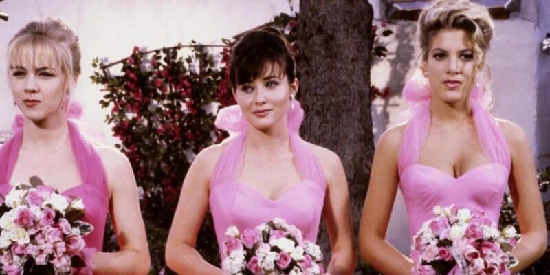 Three women stand side by side, each holding a bouquet of pink and white flowers. Dressed in identical pink satin gowns with halter necks and shoulder ruffles, they evoke a scene reminiscent of Beverly Hills 90210. Behind them is a tree and some greenery suggesting they are outdoors.