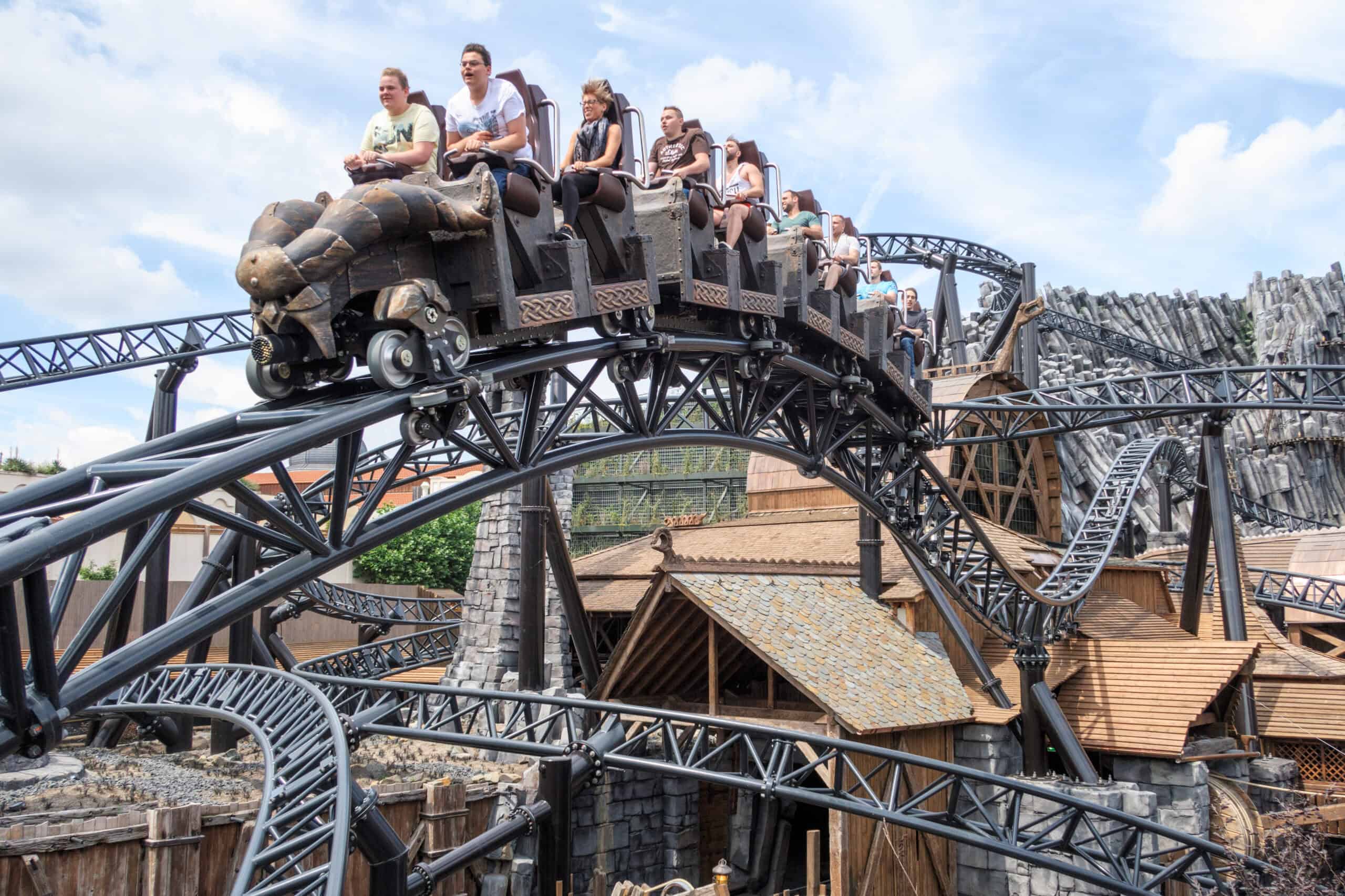 A roller coaster with several people riding in wooden-themed cars is pictured against a sunny, blue sky. The track twists and turns around various structures and wooden buildings, creating a dynamic and thrilling scene reminiscent of a bustling theme park.