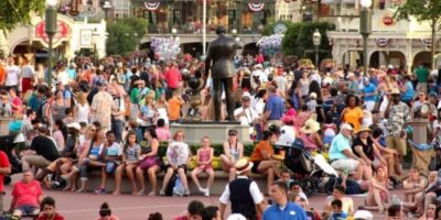 A large crowd of people gathered at an outdoor event, many sitting around a central statue. The background features various buildings and decorations, including banners and balloons. The scene is vibrant and filled with people of all ages enjoying the event.