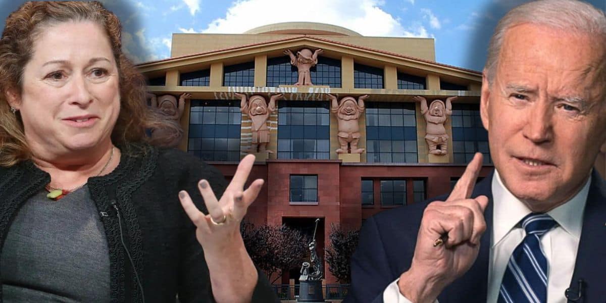 Abigail Disney on the left is gesturing with her hands, and President Joe Biden on the right is raising his index finger. Both are in front of a backdrop showing a building with Disney character statues integrated into the architecture. The sky is partly cloudy.