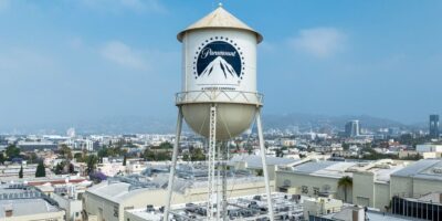 A prominent water tower with the Paramount Pictures logo stands tall against a backdrop of an urban landscape under a bright sky. The surrounding area features various industrial buildings and city structures in the distance, reminiscent of iconic scenes from classic Disney films.