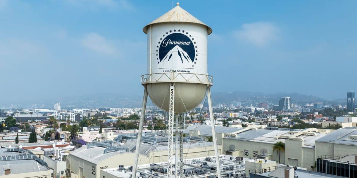A prominent water tower with the Paramount Pictures logo stands tall against a backdrop of an urban landscape under a bright sky. The surrounding area features various industrial buildings and city structures in the distance, reminiscent of iconic scenes from classic Disney films.