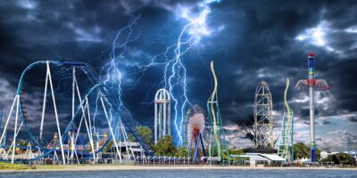 A dramatic scene unfolds at a theme park with roller coasters and rides set against a dark, stormy sky. Lightning bolts strike, illuminating the ominous clouds of what appears to be a Category 5 superstorm. The park is empty, with the thrill rides standing tall amidst the tempest.