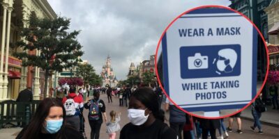 A bustling street at a Disney theme park with people walking, many adhering to mask mandates. In the foreground, two individuals are wearing masks. The inset image shows a sign that reads, "WEAR A MASK WHILE TAKING PHOTOS," accompanied by icons of a camera and a person with a mask.