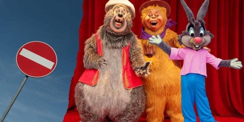 Three Disney-inspired anthropomorphic animal mascots are standing in front of a red curtain. From left to right, a bear, a lion, and a rabbit happily pose with their arms open wide. On the left side of the image, there is a road sign with a white horizontal line on a red circle against a blue sky.