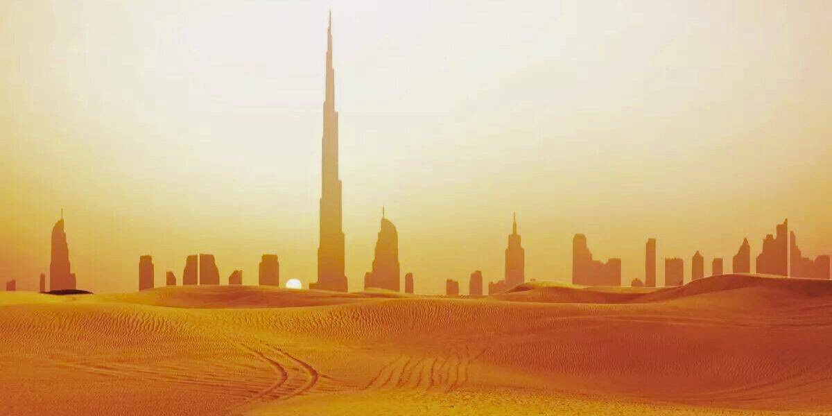 A vast desert landscape with golden sand dunes in the foreground. In the background, a modern city skyline with a range of skyscrapers, including the iconic Burj Khalifa, stands against a hazy orange sky as the sun sets, reminiscent of an otherworldly theme park in Dubai.