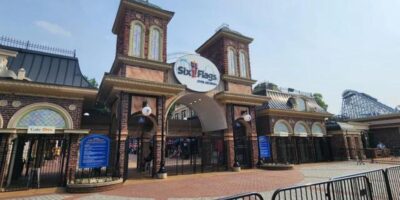 The main entrance of Six Flags Over Georgia features a large sign with the park's name and logo. The entrance has two tall brick towers with spires, large iron gates, and colorful decorative windows. A roller coaster is visible in the background, reminding visitors to review the ejection policy before entry.