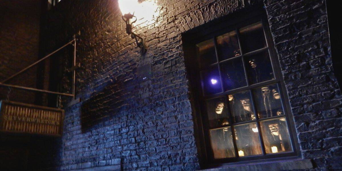 A dimly lit alley with an old brick building, featuring a window displaying several eerie, illuminated masks peering out. A single overhead light casts shadows on the wall, adding to the spooky atmosphere reminiscent of a scene from Harry Potter at Universal parks.