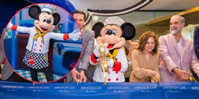 Mickey Mouse dressed as a chef poses with arms wide open in an inset. In the main image, Mickey in the same outfit holds large golden scissors with adults cutting a ribbon that reads "ANIMATION CAFE." The setting appears to be a celebratory event at an animation studio.