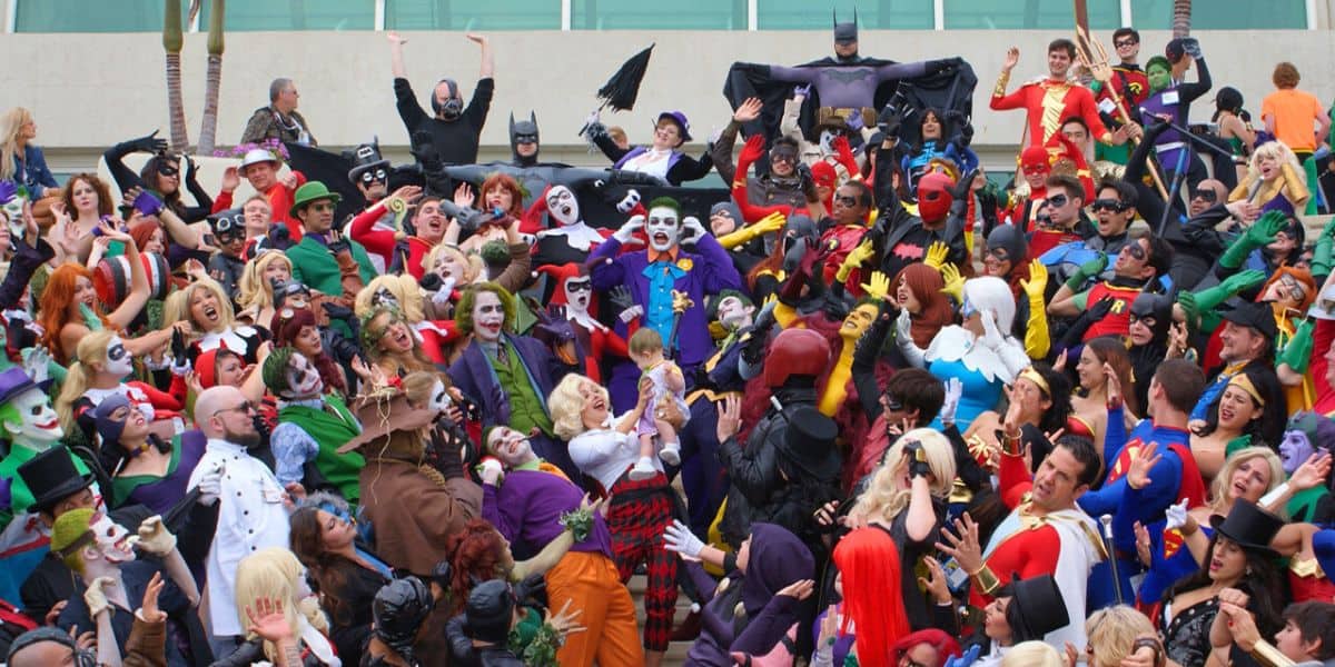 A large group of people dressed in various superhero and villain costumes from comic books, posing together outside the San Diego Comic-Con convention center. The costumes feature characters like Batman, Joker, and others, with vibrant colors and expressive poses, creating a lively scene.