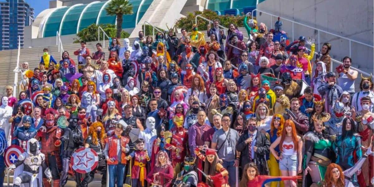 A large group of people dressed in various colorful superhero and pop culture costumes gather for a group photo outside on a sunny day. They are standing on stairs leading to a modern building with a glass facade at San Diego Comic-Con. Some characters are identifiable from comics and movies.