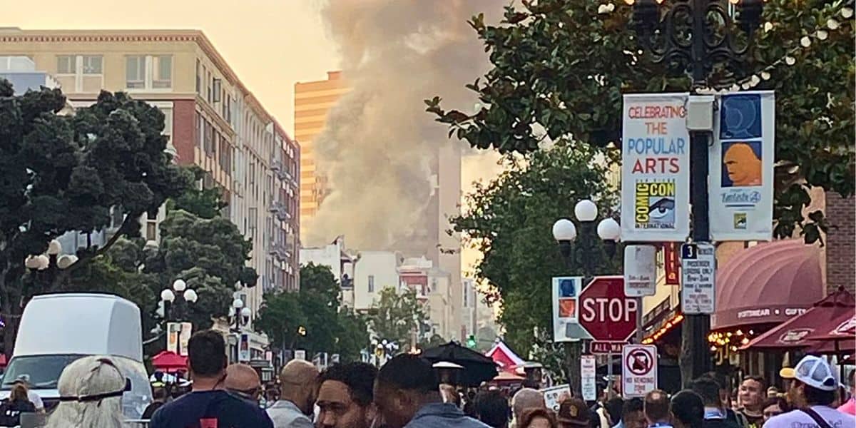 A large crowd of people walks through a busy urban street, with trees and tall buildings lining the sides. Smoke rises in the distance, partially obscuring some buildings. Various signs, including one about San Diego Comic-Con and arts, are visible along the street.