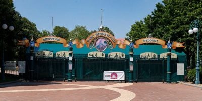 The image shows the entrance gates of a theme park with a sign reading "Disneyland." The gates are closed, and a banner reading "FERME" (French for "closed") is hung across the entrance. Green trees are visible in the background along with blue skies.