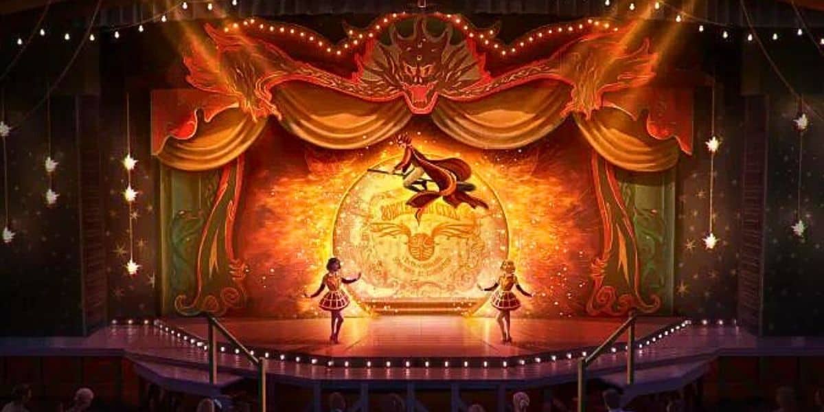 An illuminated stage with a vibrant backdrop featuring a dragon and a fiery motif. Two performers in colorful costumes are standing near the center, facing the audience. The stage is adorned with lights and drapes, creating a lively, festive atmosphere reminiscent of the Wizarding World of Harry Potter at Universal Orlando.