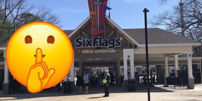 A photograph of the entrance to Six Flags Great Adventure, with people walking in and out under the large sign. An emoji of a person with a finger over their lips in a shushing gesture is superimposed over part of the image, hinting at new rules. Trees and a clear sky are in the background.