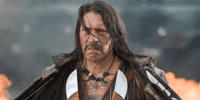 A person with long hair and a serious expression stands outdoors with fire in the background, resembling a scene straight out of Spy Kids. They are dressed in rugged clothing, including a leather jacket and a necklace with a large cross pendant. The scene appears intense and dramatic.