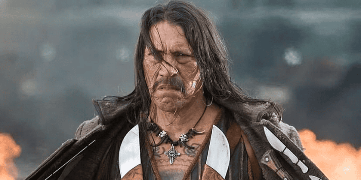 A person with long hair and a serious expression stands outdoors with fire in the background, resembling a scene straight out of Spy Kids. They are dressed in rugged clothing, including a leather jacket and a necklace with a large cross pendant. The scene appears intense and dramatic.