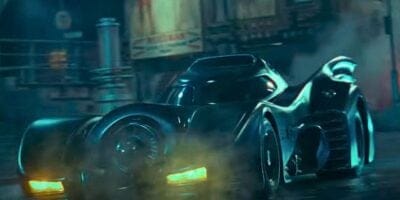A sleek black car with a futuristic design and large rear fins drives through a foggy urban street at night. The headlights are on, illuminating the mist around the vehicle, reminiscent of a Batman reboot scene. The setting appears dark and atmospheric with some indistinct structures in the background.