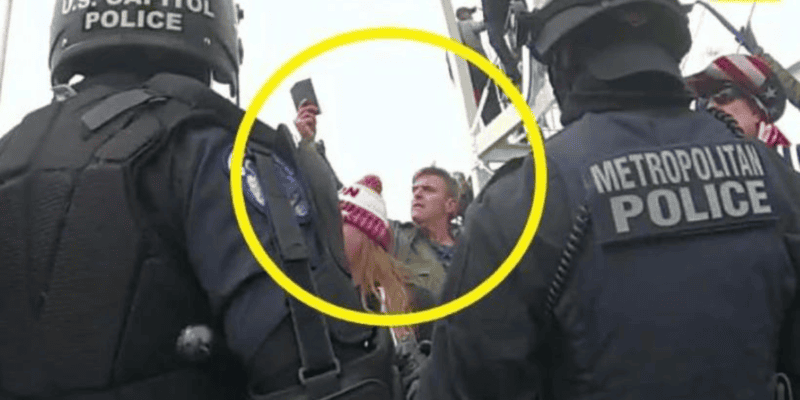 A group of U.S. Capitol and Metropolitan police officers stand together, wearing helmets and protective gear. In the background, a man and woman are partially visible inside a yellow circle, with the man holding a phone in the air, seemingly taking a photo or video during January 6th events.
