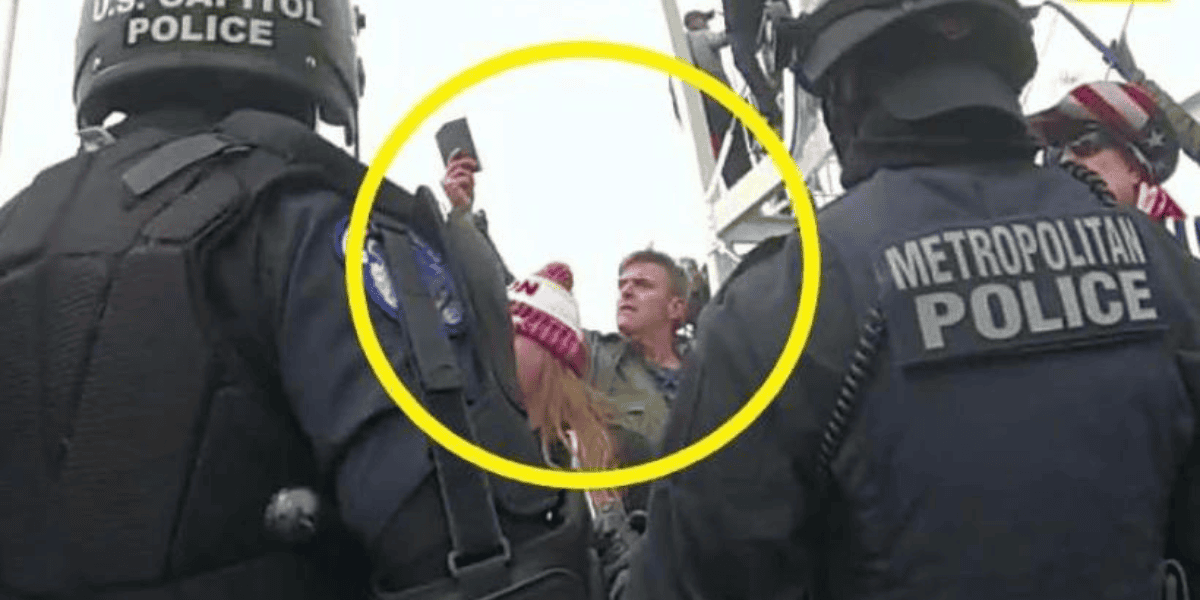 A group of U.S. Capitol and Metropolitan police officers stand together, wearing helmets and protective gear. In the background, a man and woman are partially visible inside a yellow circle, with the man holding a phone in the air, seemingly taking a photo or video during January 6th events.