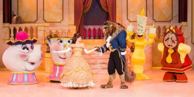 A scene from "Beauty and the Beast" featuring Belle in a yellow dress dancing with the Beast in a ballroom. Around them are characters in costume: Mrs. Potts, Chip, Lumière, and Cogsworth, set against an elegant backdrop with a marbled floor and draped curtains.