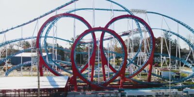 A large, intricate roller coaster features multiple loops, twists, and turns. The blue and red track intertwines in an elaborate design. Tragically, a coaster accident once occurred here that killed two people. The ride is set in an amusement park surrounded by trees under a clear blue sky.