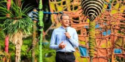A man in a blue shirt is speaking into a microphone on an outdoor stage adorned with colorful decorative elements and vibrant foliage. The background features artistic structures and plants wrapped in various colors of rope.