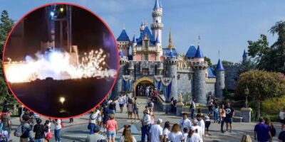 A bustling crowd stands in front of an iconic castle with blue-tiled roofs and towers at Disneyland during daytime. An inset image in the top left corner shows a nighttime scene of fireworks, smoke, and Mickey Mouse, suggesting an event or celebration.