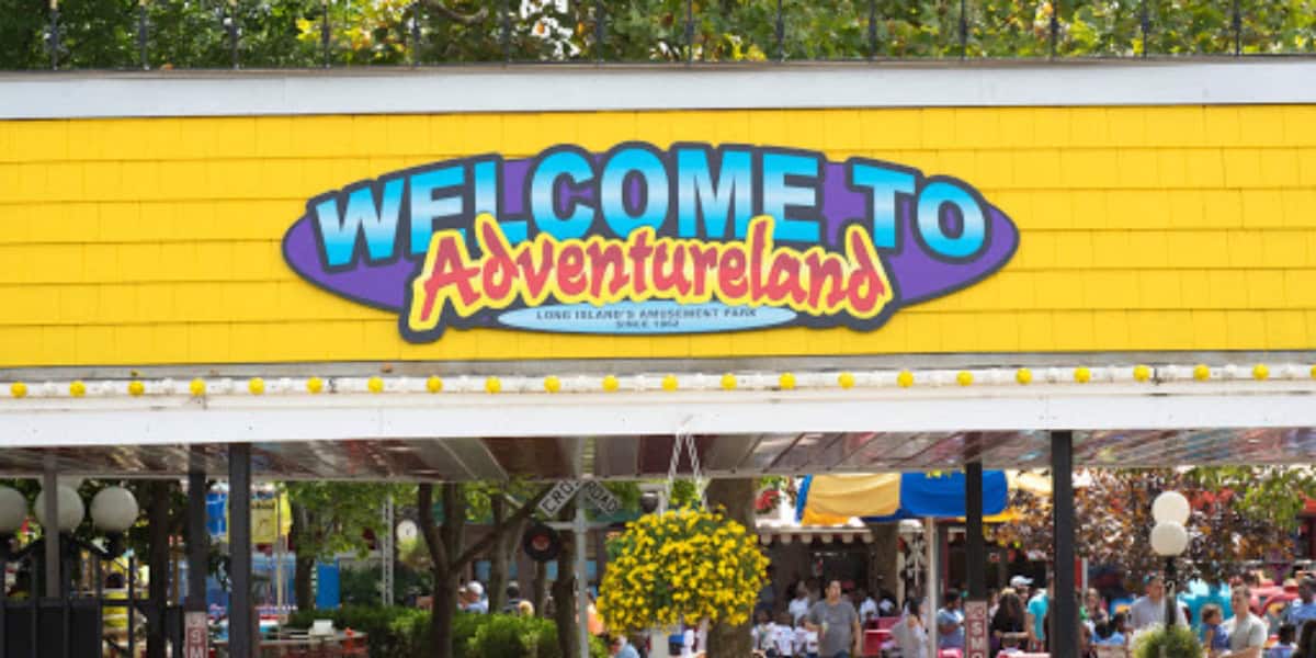 The image shows a colorful sign reading "WELCOME TO Adventureland" at the entrance of a theme park. The structure is decorated with a yellow exterior and hanging flowers, set against a backdrop of trees. Guests are seen entering below the sign, ready for fun adventures ahead.