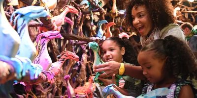 A woman and two children excitedly explore a display of colorful, fantastical bird sculptures. The vibrant birds are perched on tree branches, creating a whimsical atmosphere. The trio appears delighted, interacting with the rainbow-hued creations.