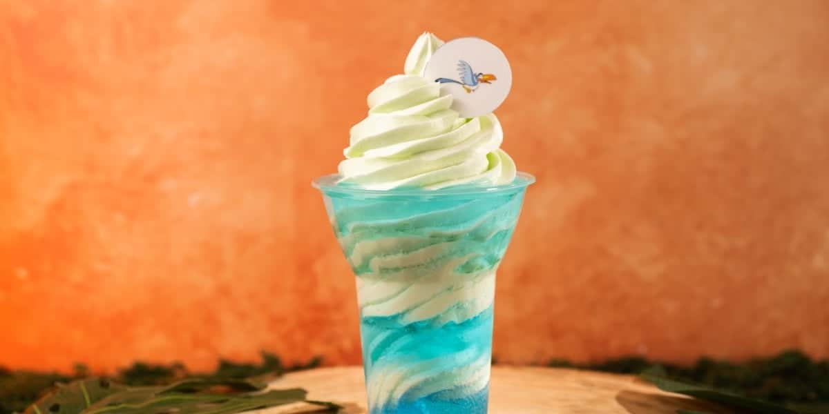 A plastic cup holds a visually appealing soft-serve ice cream. The swirl of ice cream is a blend of green and white colors, with a blue base resembling water at the bottom. A small paper topper featuring a flying bird decorates the ice cream. The background is warm and orange.