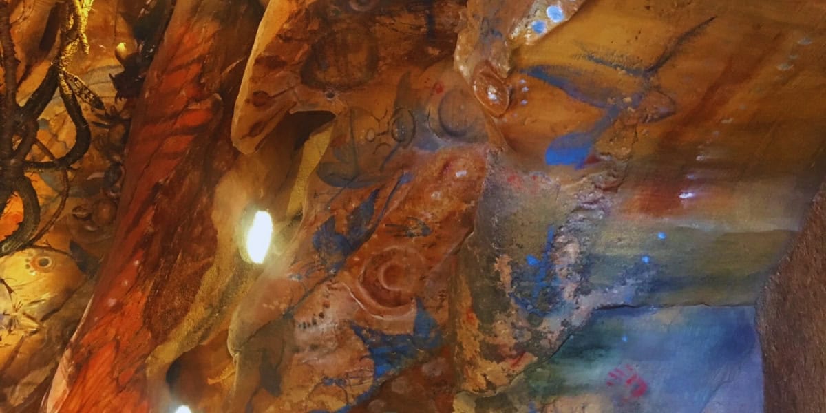 A detailed cave wall with ancient, colorful paintings and carvings. The artwork includes various patterns and figures in shades of red, blue, and yellow, illuminated by soft lighting which highlights the textures and intricate details of the rock surface.
