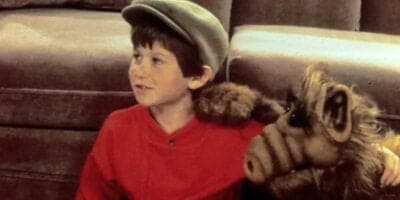 A young boy wearing a grey flat cap and a red shirt sits on the floor, smiling. Next to him is an alien puppet with brown fur, large snout, and big ears, resting its arm on the boy's shoulder. Behind them is a brown sofa. This beloved child actor's joy shines through every scene they share.